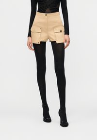 Μοντέλο που φοράει μπεζ σορτς με ψηλή μέση και μεγάλες μπροστινές τσέπες, συνδυασμένα με μαύρες καλσόν και μαύρα ψηλοτάκουνα παπούτσια.