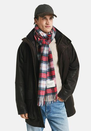 Mann trägt eine schwarze Mütze, eine dunkelbraune Lederjacke, einen cremefarbenen Pullover, blaue Jeans und einen karierten Schal in Rot, Weiß und Marineblau mit GANT-Label.