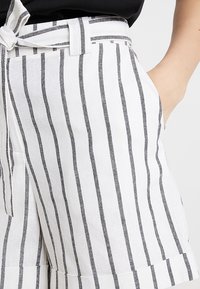 Białe lniane szorty z pionowymi czarnymi paskami, z wiązanym pasem i bocznymi kieszeniami, o luźnym kroju i gładkiej fakturze.