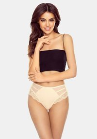 Frau mit langen braunen Haaren, die ein schwarzes, kurzes Tanktop und beigen, mit Spitze verzierten, hochgeschnittenen Unterhosen trägt, lächelt mit der Hand nah am Gesicht.