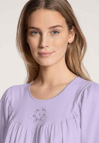 Top lavanda a maniche corte con colletto rotondo e dettagli arricciati. Presenta una leggera ricamatura floreale sulla zona del petto. Tessuto morbido.