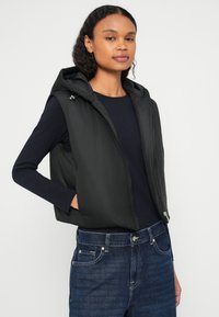 Μαύρο αμάνικο μπουφάν με επένδυση και κουκούλα, με φερμουάρ και πλαϊνές τσέπες, φορεμένο πάνω από μακρυμάνικη μπλούζα ναυτικού χρώματος και τζιν παντελόνι.