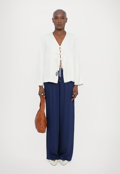 White blouse with lace-up front, paired with flowing dark blue trousers. Accessories include a woven brown bag and cream pointed shoes.