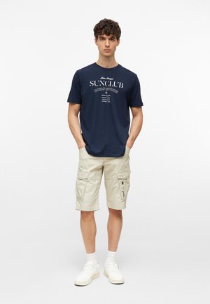 Jonge man draagt een marineblauw grafisch T-shirt, beige cargoshorts, witte sokken en witte sneakers, staand met handen in de zakken.