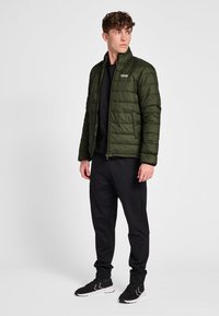 Grøn polstret jakke med høj krave, lynlås foran og vandret quiltning. Kombineret med sorte joggingbukser og sorte sneakers.