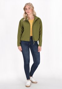 Alie žalios spalvos zip-up striukė su apykakle, derinama su geltona ir balta dryžuota marškinėlių, tamsiomis džinsinėmis kelnėmis ir baltais sportbačiais.