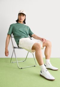 T-shirt en coton vert avec un petit logo, shorts blancs, baskets blanches avec semelles marron, assis sur une chaise en métal contre un fond uni.