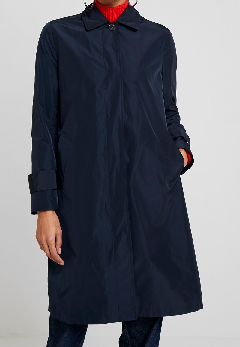Granatowy płaszcz przeciwdeszczowy wykonany z gładkiego, lekkiego materiału. Posiada kołnierz, zapięcie na guziki oraz boczne kieszenie. Długie rękawy z mankietami.