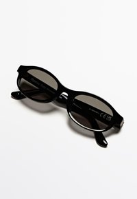 Occhiali da sole neri con forma arrotondata, dotati di lenti scure e montatura in plastica liscia. Incisione del marchio ben visibile sulla parte interna delle aste.