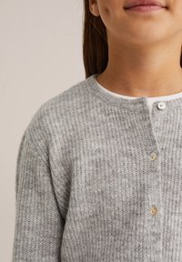 Pelēka adīta jaka ar ribotas struktūras virsmu, apaļu kakla izgriezumu un trim pogām aizdarei. Audums izskatās mīksts un silts.