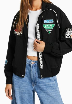 Femme portant une veste bomber noire avec des patchs colorés, un t-shirt court blanc et un jean taille haute bleu clair, tenant la fermeture éclair de la veste.