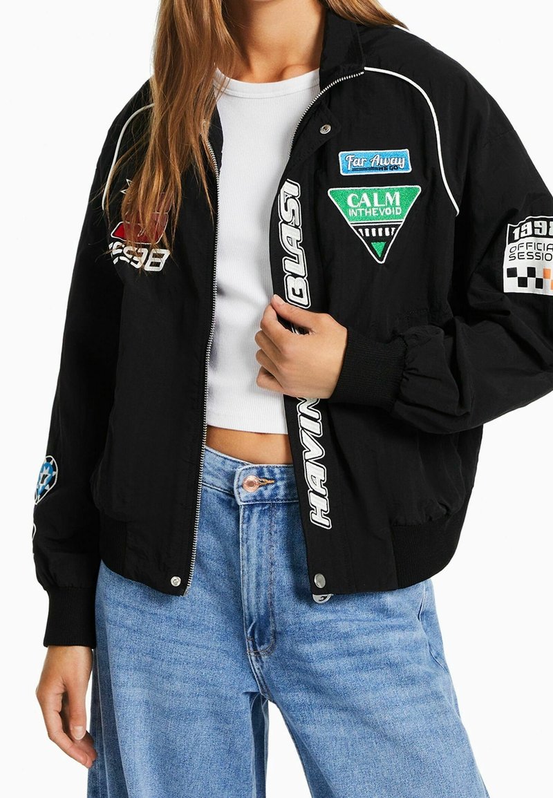 Femme portant une veste bomber noire avec des patchs colorés, un t-shirt court blanc et un jean taille haute bleu clair, tenant la fermeture éclair de la veste.