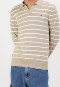 Pull-over en maille beige et blanche à rayures avec un col en V et un col. Présente un ourlet côtelé et un petit logo sur la poitrine. Coupe décontractée.