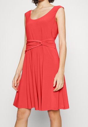 Femme portant une robe rouge sans manches avec un détail de nœud torsadé à la taille, debout devant un fond clair uni.
