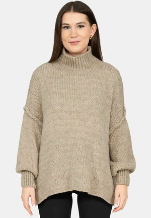 Kobieta ubrana w luźny, beżowy, gruby sweter z wysokim kołnierzem typu golf, z balonowymi rękawami oraz czarne spodnie, na jednolitym tle.