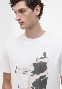 T-shirt de algodão branca com uma grande estampa de uma vaca em preto e branco. Mangas curtas e um design de gola redonda.