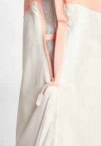 Tessuto beige chiaro con una sottile trama, caratterizzato da una zip rosa e una fodera in rete. Include un dettaglio a toggle sul tirazip.