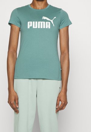 Osoba ubrana w turkusowy t-shirt Puma z białym logo i jasnoniebieskimi spodniami stojąca na jednolitym tle.