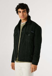 Joven vestido con una chaqueta de denin de cuello sherpa negra sobre un suéter verde y pantalones blancos, de pie contra un fondo claro y liso.