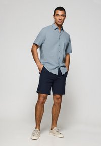 Camicia a maniche corte botton-down azzurro chiaro, pantaloni corti blu navy e sneakers beige. Il tessuto appare liscio con una vestibilità rilassata e dettagli sottili.