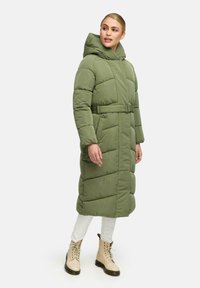Cappotto lungo e voluminoso di colore verde oliva con cappuccio, vita legata, texture trapuntata e tasche laterali, abbinato a stivali color crema con lacci.