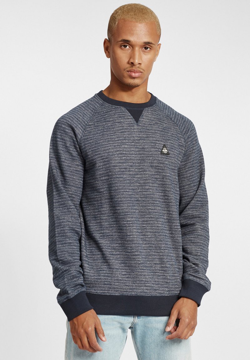 Sötétkék pulóver texturált kötésse, raglánujjakkal és bordás mandzsettákkal. A mellkasán egy kis fekete háromszög logo található.