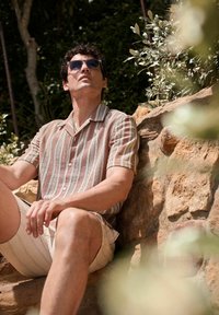 Kurzärmliges, gestreiftes Hemd in Beige- und Rosttönen, mit Knopfleiste, kombiniert mit beigen Shorts, sitzt auf einer Steinoberfläche im Freien.