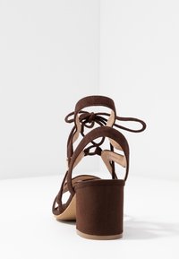Sandal de tacón alto en ante marrón con un chunky block heel, parte trasera abierta y diseño de tiras en cross, con acentos atados sobre el tacón.