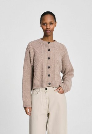 Naine, kellel on tagasikeeratud juuksed, kannab beeži värvi nööbitud soonikkoes kardigani ja heleda beeži kõrge vöökohaga pükse, seisab ühe käega taskus.