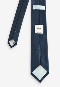 Cravat texturat de culoare bleumarin cu un capăt ascuțit. Prezintă o căptușeală interioară de culoare albastru deschis cu modele abstracte și un etichetă cu inscripția „NEXT TAILORING.”