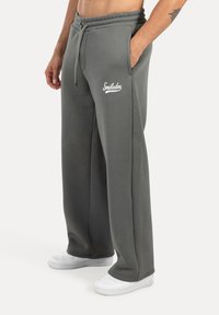 Pantalones de chándal grises con un corte relajado, que cuentan con una cinturilla con cordón y bolsillos laterales. Logo "Smiledox" en blanco en el muslo izquierdo. Zapatillas blancas.