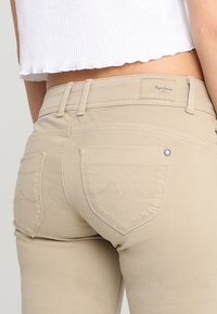 Bežové elastické kalhoty s zadními kapsami a jemnými švy. Mají úzký pas a hladký povrch. Viditelný štítek značky.