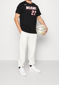 Czarna koszulka Nike z napisem "MIAMI" i "22" w czerwonych literach, białe dresy i białe sneakersy z różowymi akcentami, trzymający piłkę do koszykówki.