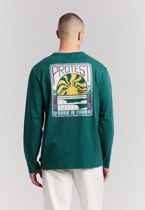 Mann mit kurzen Haaren trägt ein grünes Langarmshirt mit Grafik von Sonne, Wellen und den Texten „PROTEST“ und „MAKE IT EASY“ auf dem Rücken.
