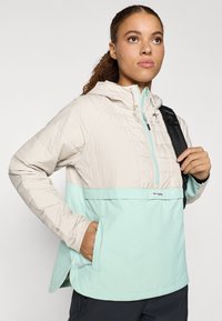 Lahka jakna s kapuco, ki ima kremasto bež zgornji del in mentolno zelen spodnji del. Vključuje sprednji žep in nastavljive vrvice.