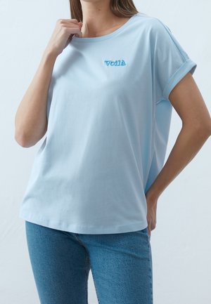 Vrouw draagt een lichtblauw shirt met korte mouwen met "Voilà" geborduurd op de linkerborst, gecombineerd met een blauwe spijkerbroek, tegen een effen achtergrond.