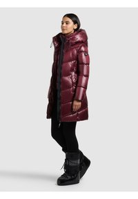 Sieviete, kura ir tērpusies garā spīdīgā bordo krāsas pūkainajā mētelī ar kapuci, melnās biksēs un melnos sniega zābakos, stāv pret vienkāršu fonu.