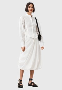 Biała koszula z długim rękawem, z marszczonymi bokami, dopasowanym korpusem i zapięciem na guziki, połączona z zwiewną białą spódnicą i czarnymi sandałami na paski.