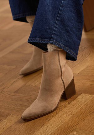 Beige suede ankle boots with block heels worn with beige socks and blue denim jeans on a wooden parquet floor.