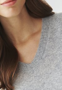 Grå V-hals sweater lavet af blødt materiale, med ribbet halsudskæring og subtil tekstur. Håret er delvist synligt på venstre side.