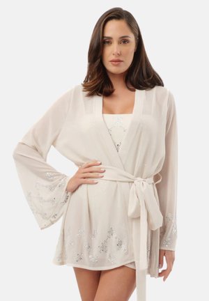 Donna con capelli castani di media lunghezza che indossa un accappatoio beige chiaro con motivi floreali argento e una cintura legata in vita, posando con una mano sul fianco.