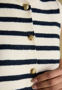 Wit en marineblauw gestreepte gebreide top met gouden ronde knopen, met een zachte textuur en een getailleerd ontwerp. Hand rustend op het kledingstuk.