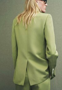 Maatwerk groene blazer met een subtiele textuur, enkele rugsplit en knoopaccenten op smalle mouwen. Ontworpen in een getailleerde snit.