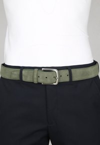Groene suede riem met een zilveren metalen gesp en meerdere gaatjes voor verstelling, gedragen over donkere broek. Zachte textuur en eenvoudig ontwerp.