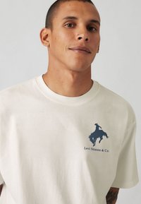 Kortærmet creme t-shirt lavet af bomuld, med et mørkeblåt cowboy-design og teksten "Levi Strauss & Co." på venstre brystområde.