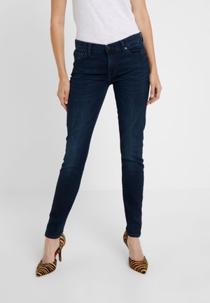 Femme portant un jean slim bleu foncé et des escarpins à rayures tigre, debout devant un fond uni clair.