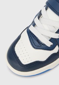 Bielo-modrá teniska so syntetickou kožou s textúrou, okrúhlou špičkou, perforovanou prednou časťou a bielymi šnúrkami. Modrá gumová podrážka.