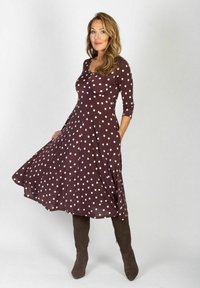 Brązowa sukienka w kropki z rozkloszowanym dołem, okrągłym dekoldem i rękawami do trzech czwartych. Modelka w brązowych kozakach do kolan.