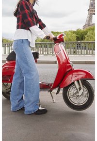 Röd vintage scooter med kromdetaljer och rund strålkastare, parkerad på en trottoar med en person som b är blå vida jeans och en rutiga topp.