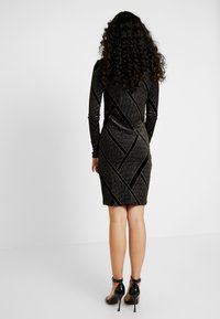 Svart bodycon klänning med långa ärmar, texturerat tyg, diagonala metallicränder och en figurnära siluett, kombinerad med svarta högklackade skor.